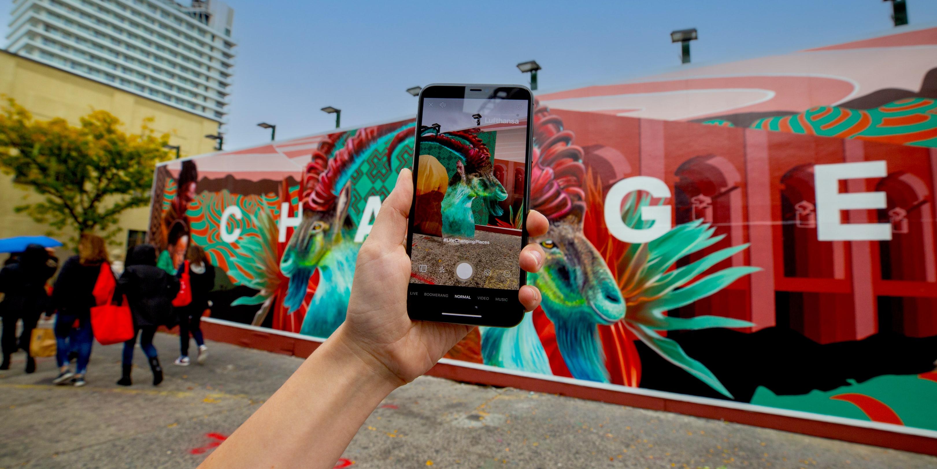 A hand holds a smartphone capturing a vibrant AR mural featuring colorful animals, while people stroll by in the urban setting.