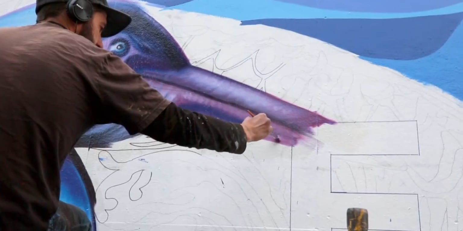 Ein Mann mit Kopfhörern malt ein lebendiges Wandgemälde auf eine weiße Wand.