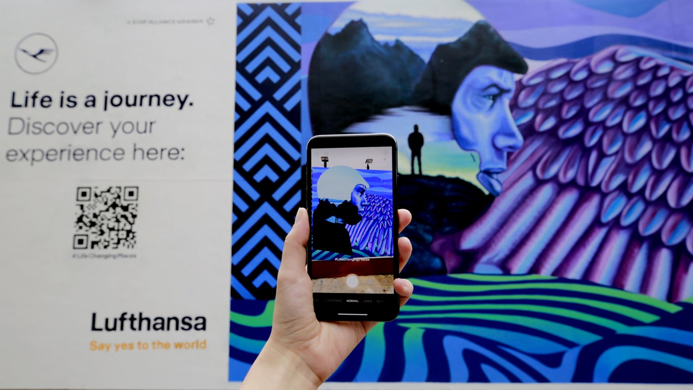 Eine Person hält ein Smartphone, das ein lebhaftes AR Mural zeigt, das Themen des Reisens und der Erkundung illustriert, begleitet von einer Lufthansa-Anzeige, die die Reise des Lebens hervorhebt.