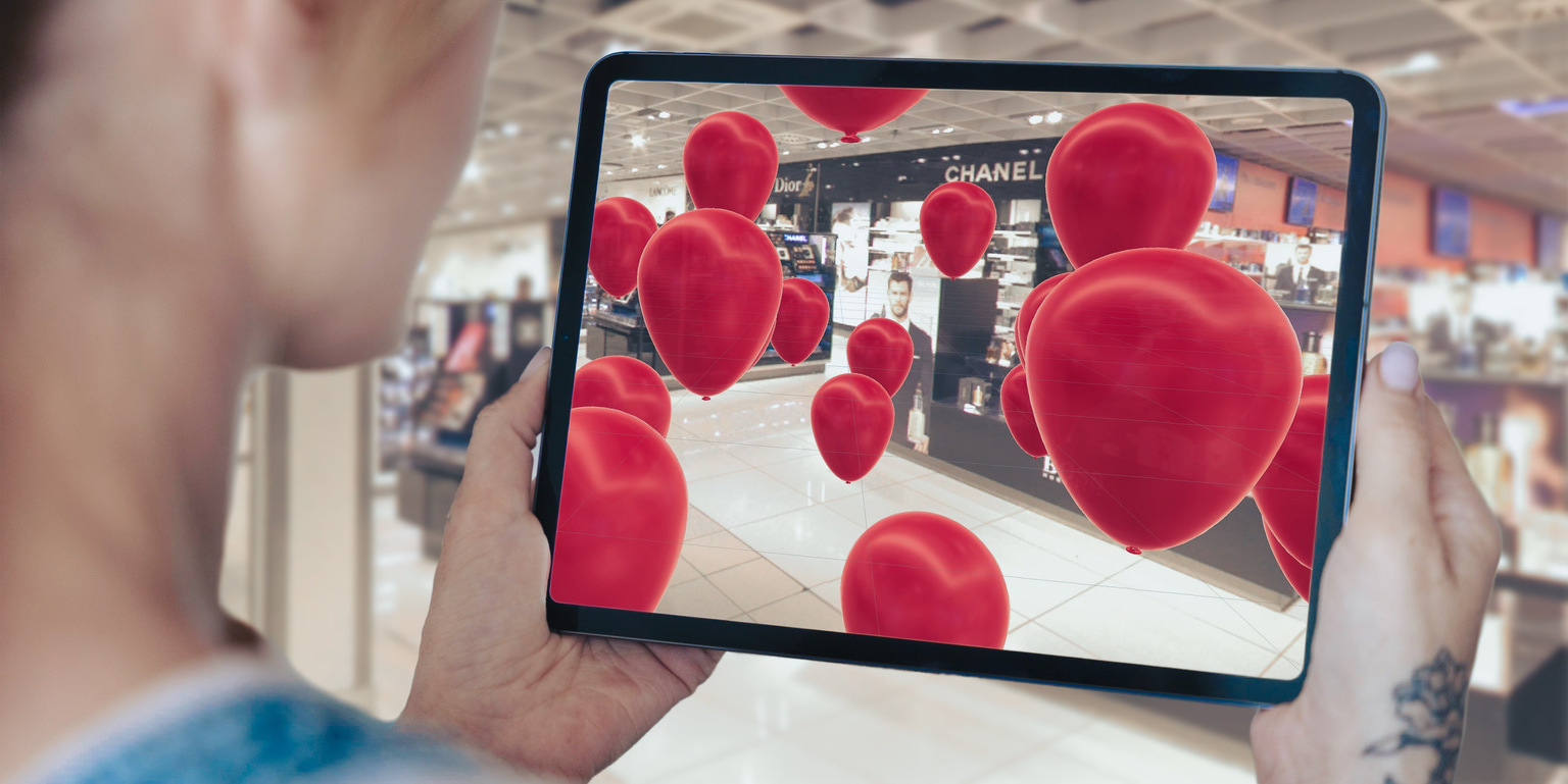 A person is using a tablet to view an augmented reality scene filled with floating red hearts in a shopping mall. 