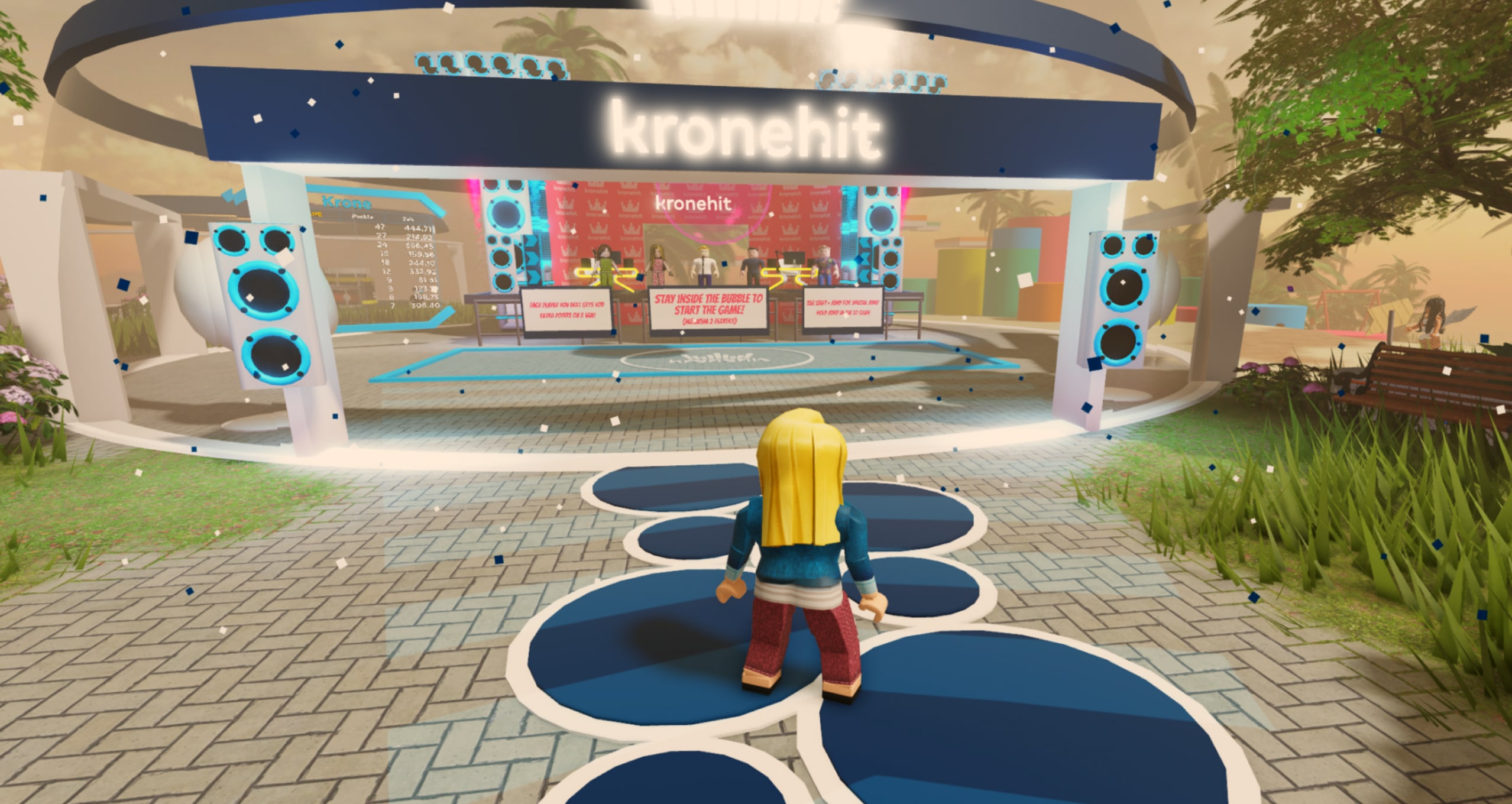 A Roblox player stands in front of a vibrant gaming stage decorated with large speakers and colorful banners.
