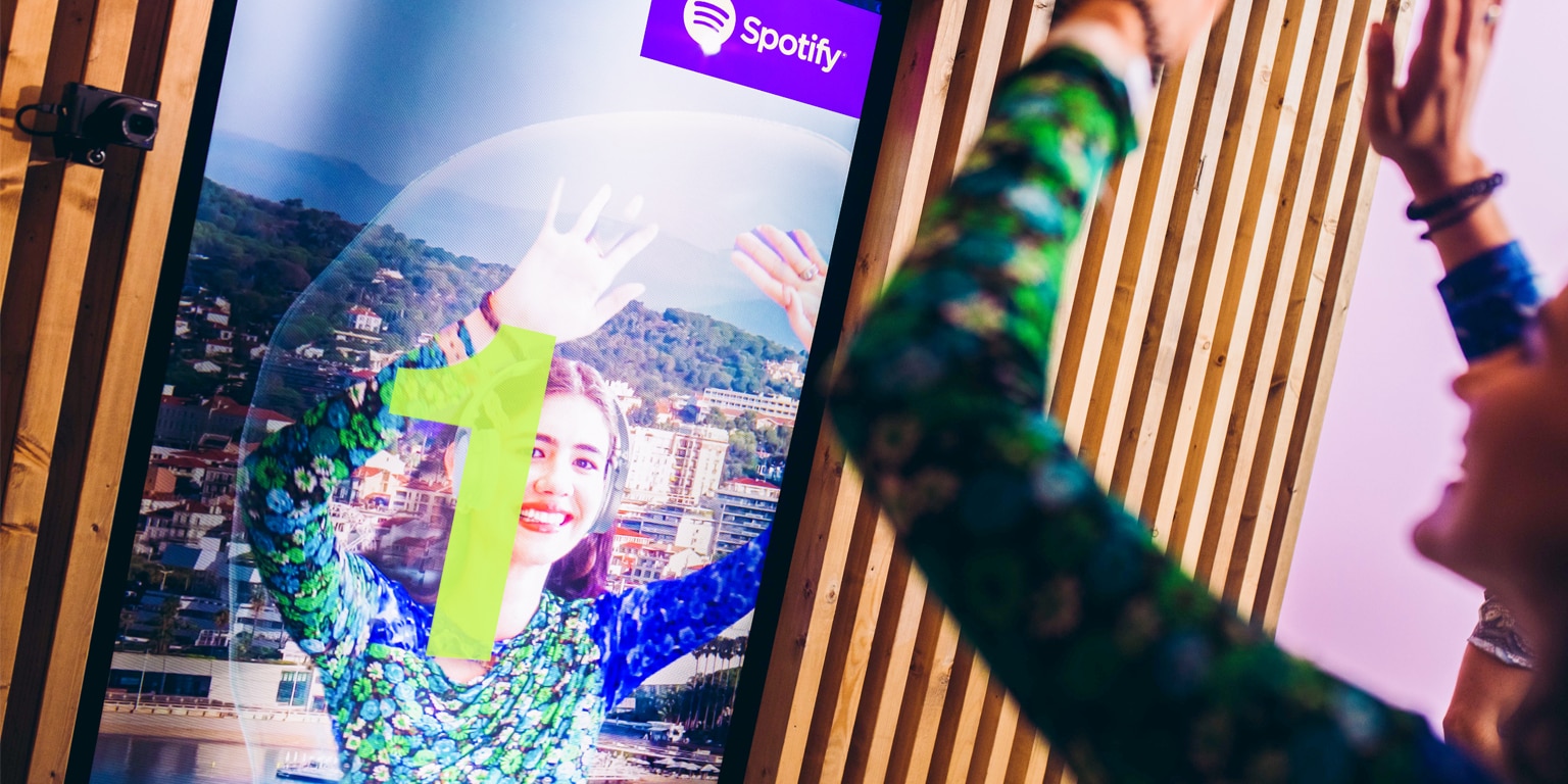 A person stands in front of a colorful digital display featuring Spotify branding, playfully interacting with the screen.